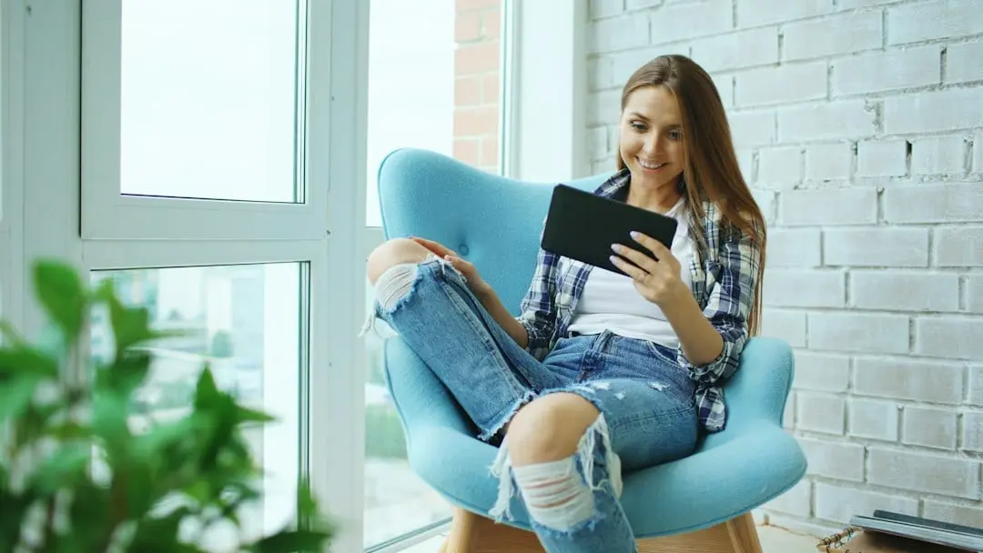 A smiling woman in a blue armchair by a window holding a tablet, as when signing in to Flixaria in a player app