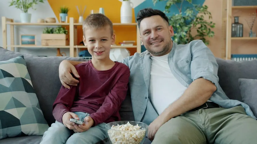 A father and his young son smiling on a grey sofa with a bowl of popcorn, settling in to watch TV with Flixaria