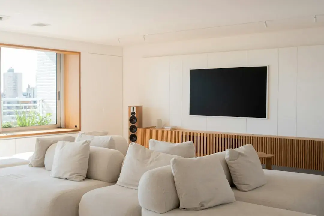 A bright living room with a wall-mounted TV above a wooden media unit, a large cream sectional sofa and a window onto the city